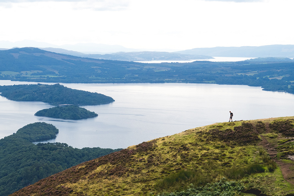 Conic Hill. A tinyfigure of a person stands on the side of a hill in front of a wide expanse of water.