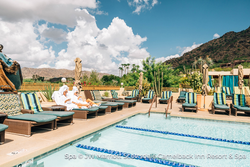 Pool at the Camelback Inn, in PHoenix, Arizona. Camelback Mountains are in the background while two people in fluffy white robes and towels wrapped around their heads sit on sun-loungers by the poolside.