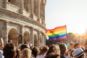 a Pride flag is waved above a crowd next to the Colosseum in Rome, Italy, at sunset.